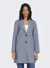 Kobieta w jasnoszarym płaszczu do kolan z dwoma czarnymi guzikami, na białym topie i niebieskich dżinsach, stojąca na jednolitym tle.