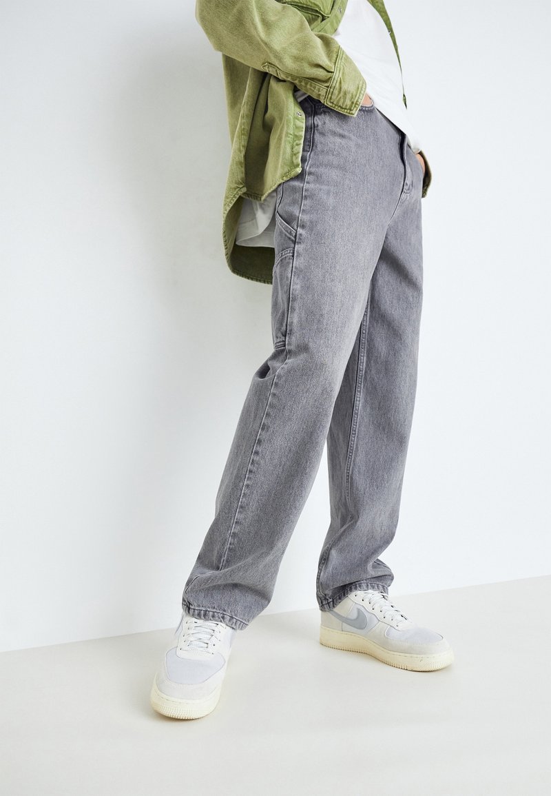 Pantalones de mezclilla gris con un corte relajado, que cuentan con cinco bolsillos. Combinados con zapatillas blancas y una chaqueta verde oliva.