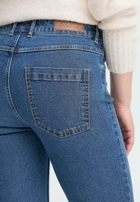 Artimas žmogaus plano atvaizdas, kur matomi mėlyni džinsai su užpakaliniu kišeniu ir odinė prekės ženklo etiketė, kartu su matoma kremine megztinio rankove.