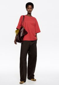 Camiseta de algodón roja de corte oversize, combinada con pantalones anchos oscuros. Accesorios con un bolso de piel marrón que presenta un acento de piel amarilla.