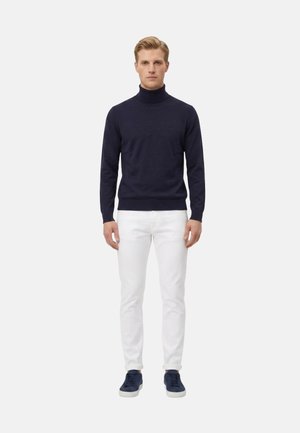 Uomo in piedi di fronte, indossa un maglione dolcevita blu navy, pantaloni bianchi e scarpe da ginnastica blu navy su uno sfondo grigio uniforme.