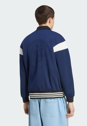 Giovane uomo che indossa una giacca blu navy con orlo e maniche a righe nere e bianche, visto di spalle, abbinata a pantaloni azzurri con tasche posteriori.