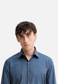 Camicia blu con bottoni e motivo a trama. La camicia presenta un colletto, maniche lunghe e una vestibilità slim, indossata da una persona con capelli ricci.