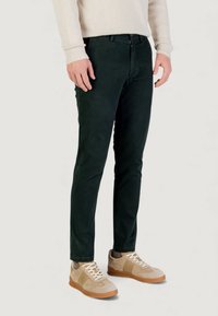 Pantalones de corte slim de color verde oscuro hechos de tela suave, con cremallera frontal y bolsillos, combinados con zapatillas beige y un suéter ligero.