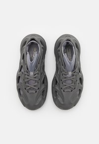 Šedé sportovní boty s tvarovanou vnější skořepinou, síťovanými akcenty a šněrováním. Mají jedinečný výřez pro ventilaci a flexibilitu.
