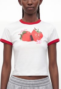 Γυναίκα που φοράει λευκό cropped t-shirt με κόκκινο περίγραμμα και γραφικό δύο φραουλών και το κείμενο "Jordan 23" στο μπροστινό μέρος.