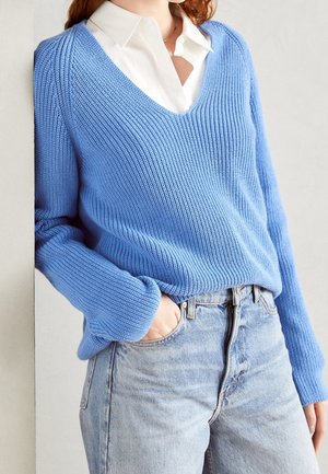 Vrouw in een lichtblauwe ribgebreide trui over een witte blouse, met haar hand in de zak van een hooggetailleerde lichtblauwe jeans.