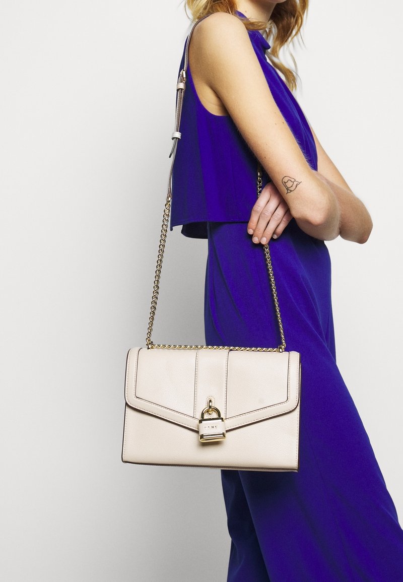 Borsa a spalla in pelle crema con tracolla in catena dorata, caratterizzata da una chiusura a scatto e dettagli di design geometrico. Tessuto liscio, forma strutturata.