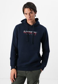 Marineblauwe fleece hoodie met een voorste kenguruzak en een trekkoord. Voorzien van een wit en rood "tommy jeans" logo op de borst.