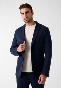 Marineblauwe blazer van lichtgewicht stof met een getailleerde pasvorm. Voorzien van ingesneden revers, twee knopen en voorzakken. Gecombineerd met een crèmekleurig overhemd.