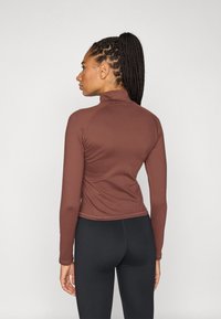 Καφέ μακρυμάνικη μπλούζα με ψηλό γιακά, εφαρμοστή γραμμή και λεία υφή, συνδυασμένη με μαύρο κολάν. Η όψη από πίσω δείχνει λεπτομέρειες με ραφές.