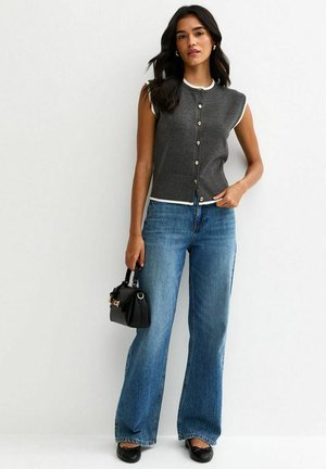 Donna che indossa un top grigio senza maniche con bottoni e bordi bianchi, jeans blu, ballerine nere, tiene una piccola borsa nera, in piedi contro un muro bianco.