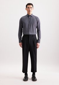 Grijs geruite shirt met knopen, gecombineerd met zwarte broek met hoge taille. De broek heeft plooien en opgerolde manchetten; de schoenen zijn van zwart leer.