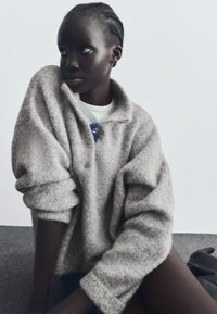 Prevelik, sivi vuneni pulover s ovratnikom i zatvaranjem na gumbe; ima teksturiranu površinu i zavrtnje rukava. Pozadina je neutralna.