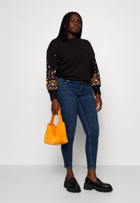 Sieviete, tērpusies melnā džemperī ar ziedu piedurknēm, zilo džinsu biksēs, melnās masīvās loafer kurpēs, tur spožu oranžu rokassomu, stāvot pret baltu fona.