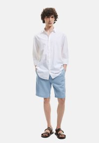 Camisa blanca de botones, pantalones cortos azul claro, sandalias negras. Atuendo casual con un corte relajado y mangas cortas. Textura de tela suave.
