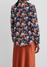 Camicia floreale blu navy con maniche lunghe, caratterizzata da fiori arancioni e bianchi. Il tessuto è liscio, con orlo arrotondato e polsini con bottone.