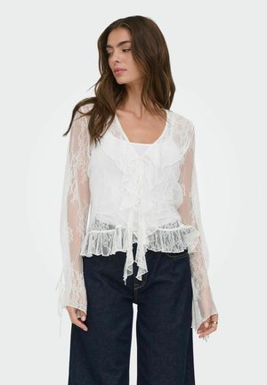 Femme portant un chemisier blanc en dentelle à manches longues avec des volants et un jean bleu foncé, regardant vers sa gauche sur un fond uni.