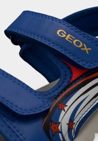 Μπλε λουρί παιδικού σανδαλιού με κίτρινο κείμενο «GEOX» και διακοσμητικά κόκκινα αστέρια με άσπρες και μαύρες ρίγες που ακολουθούν στο πλάι.