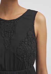 Svart klänning med intrikata svarta spetsdetaljer på axlarna och i ringningen, med en mjuk, flödande tygtextur och ärmlös design.