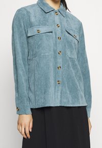 Ljusblå kordjakett med krage, två bröstfickor och bruna knappar; textil med vertikala ribbor.