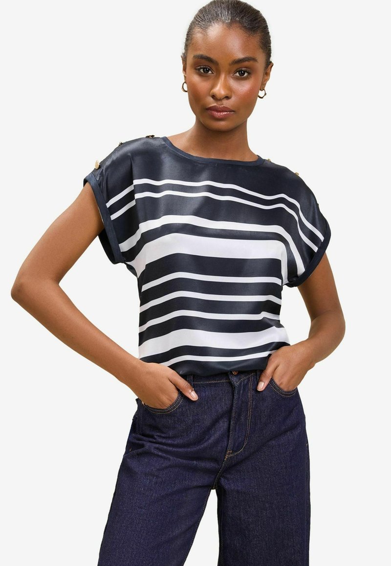 Marineblå og hvidstripet bluse i glat stof, med korte ærmer og guld hardware-accenter på skuldrene. Iført med mørk denimjeans.