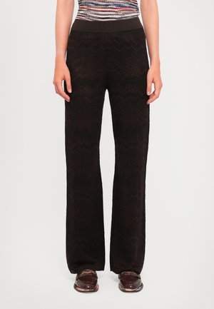 Donna con pantaloni a gamba larga neri e strutturati, top multicolore a fantasia e mocassini marroni scuri in piedi su uno sfondo bianco.