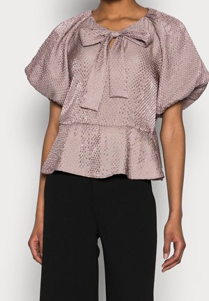 Błyszcząca, fakturowana bluzka w kolorze mauve z bufkami na krótkich rękawach, wiązana kokardą z przodu przy dekolcie i z baskinką, noszona z czarnymi spodniami.