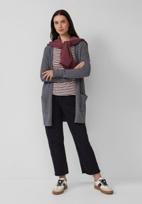 Une femme se tient avec les bras croisés, portant un cardigan gris sur une chemise rayée, un pantalon noir coupé, des baskets blanches et un sweat à capuche bordeaux drapé sur les épaules.