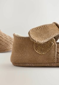Chaussures pour bébé en daim marron avec une doublure douce, dotées d'une sangle en velcro et d'accents texturés sur le côté. Forme arrondie avec un bord cousu.