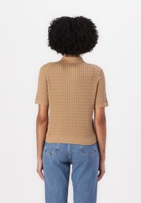 Maglione a maniche corte, a coste, in beige con un motivo testurizzato. Presenta un colletto rotondo e un orlo aderente. Indossato con jeans azzurri chiari.