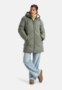 Doudoune vert olive, avec une capuche, design matelassé, fermeture éclair frontale et poches latérales. Associée à un jean large bleu clair et des bottes marron.
