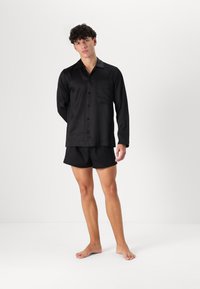 Svart satin pyjamas-set bestående av en långärmad skjorta med knappar och bröstficka samt matchande shorts, slät textur och klassisk passform.