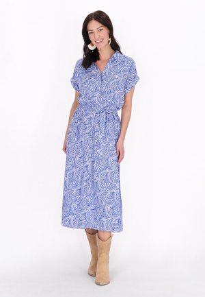 Maxikleid mit blauem Paisleymuster, kurzen Ärmeln und taillierter Taille. Getragen mit beigen Ankle Boots. Glatte Stofftextur.