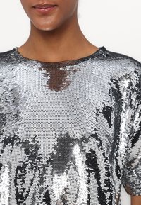 Silvrig paljettblus med lös passform och vid halsringning. Den texturerade ytan reflekterar ljus, med ett nätliknande mönster under.