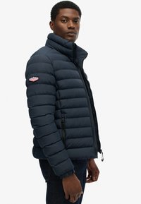Marineblå pufferjakke med vandret syninger, høj krave, lynlåslommer og et logo-patch på venstre ærme. Struktureret stof og en tætsiddende pasform.
