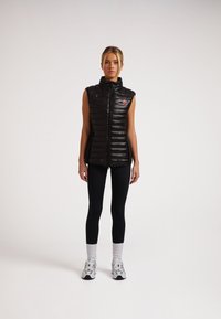 Schwarze ärmellose Steppweste mit hohem Kragen, gestepptem Design, durchgehendem Reißverschluss und einem roten Logo-Patch. Kombiniert mit schwarzen Leggings und Turnschuhen.