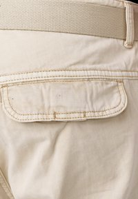 Helle beigefarbene Hosen mit einer flachen Gesäßtasche, kontrastierenden Nähten und einem strukturierten Bund. Der Stoff wirkt weich und glatt.