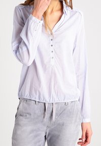 Chemise bleu clair à boutons avec col, manches longues, ourlet élastique et petits pois blancs subtils. Associée à un pantalon gris clair.