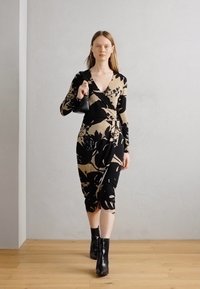 Pitkähihaista kietaisumekkoa beige-värissä mustalla kukkakuviolla, jossa on V-mallinen kaula-aukko ja drapattu muotoilu. Mallilla on päällään mustat nilkkurit ja hän pitää laukkua.