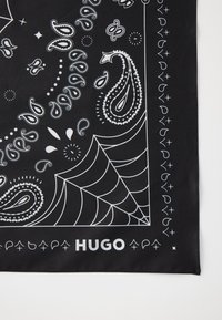 Fekete bandana fehér paisley és geometrikus mintákkal, az alábbi sarokban "HUGO" felirattal, fehér színben. Sima szövet textúra.