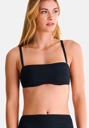 Jeune femme aux cheveux blonds portant un haut de bikini bandeau noir texturé avec de fines bretelles, tournée légèrement vers la droite.