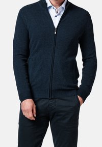 Tmavě modrý kardigan na zip z texturované látky, s vysokým límcem, žebrovanými manžetami a dvěma bočními kapsami. Nosit přes bílou košili s límečkem.