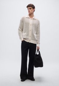 Πλεκτή ζακέτα σε εκρού με υφασμένο μοτίβο, μεγάλο γιακά και κουμπιά; συνδυασμένη με φαρδιά σκούρα τζιν παντελόνια και μαύρη τσάντα.