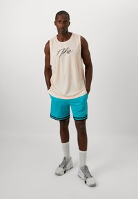 Helles Pfirsichfarbener, ärmelloser Mesh-Top mit schwarzer Schrift, kombiniert mit türkisfarbenen Shorts mit schwarzen Streifen. Getragen mit weißen Socken und Turnschuhen.
