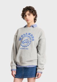 Sudadera gris con gráfico azul "PARQUE NACIONAL DE BIENESTAR", cuello redondo, corte oversized, superpuesta sobre una camisa de rayas azul y blanca, falda de mezclilla.