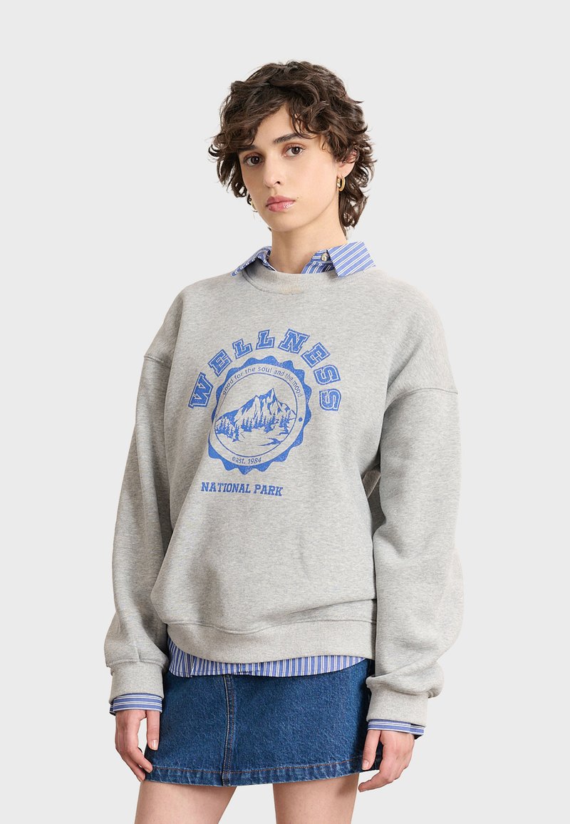 Sudadera gris con gráfico azul "PARQUE NACIONAL DE BIENESTAR", cuello redondo, corte oversized, superpuesta sobre una camisa de rayas azul y blanca, falda de mezclilla.