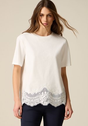 Mujer vestida con una camiseta blanca de manga corta con ribete de encaje floral en el borde inferior y pantalones oscuros, posando frente a un fondo neutro.