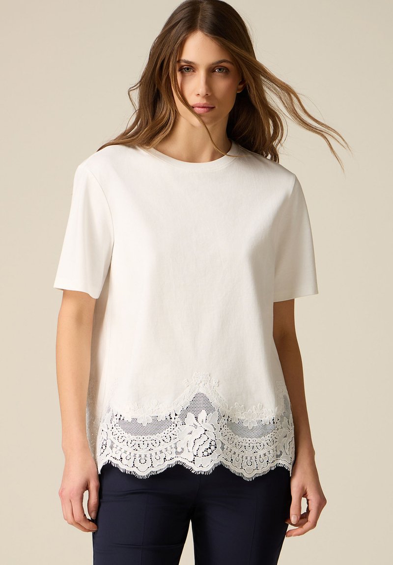 Nainen, joka pukeutunut valkoiseen lyhythihaiseen yläosaan, jossa on kukkainen pitsireunus helmassa, ja tummiin housuihin, seisoo neutraalia taustaa vasten.