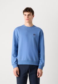 Light blue sweatshirt made of soft fabric, features a crew neck, ribbed cuffs, and a small logo patch on the left chest. Simple design.